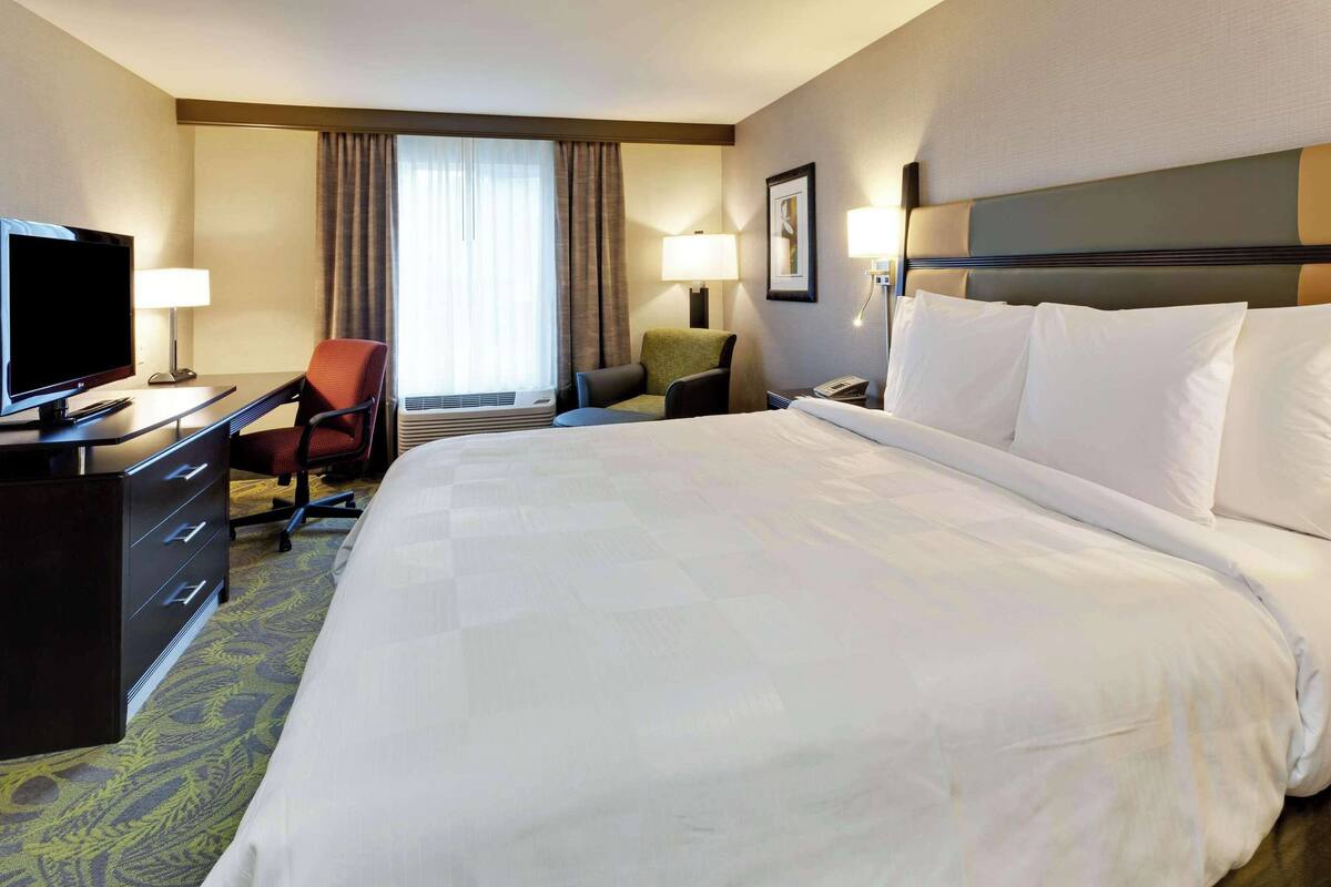 A hotel room with a large bed, a desk with a television, a chair, and a window with curtains.