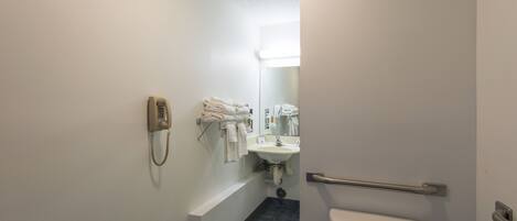 Un baño con inodoro, lavamanos y espejo. Hay un teléfono en la pared y toallas colgadas en un perchero.