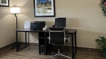 A hotel room with a desk, chair, printer, lamp, framed picture, and a potted plant.
