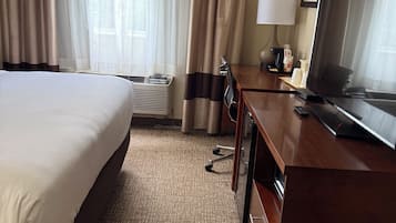 Habitación de hotel con una cama grande, un escritorio con televisor, una lámpara y un ventanal con cortinas.