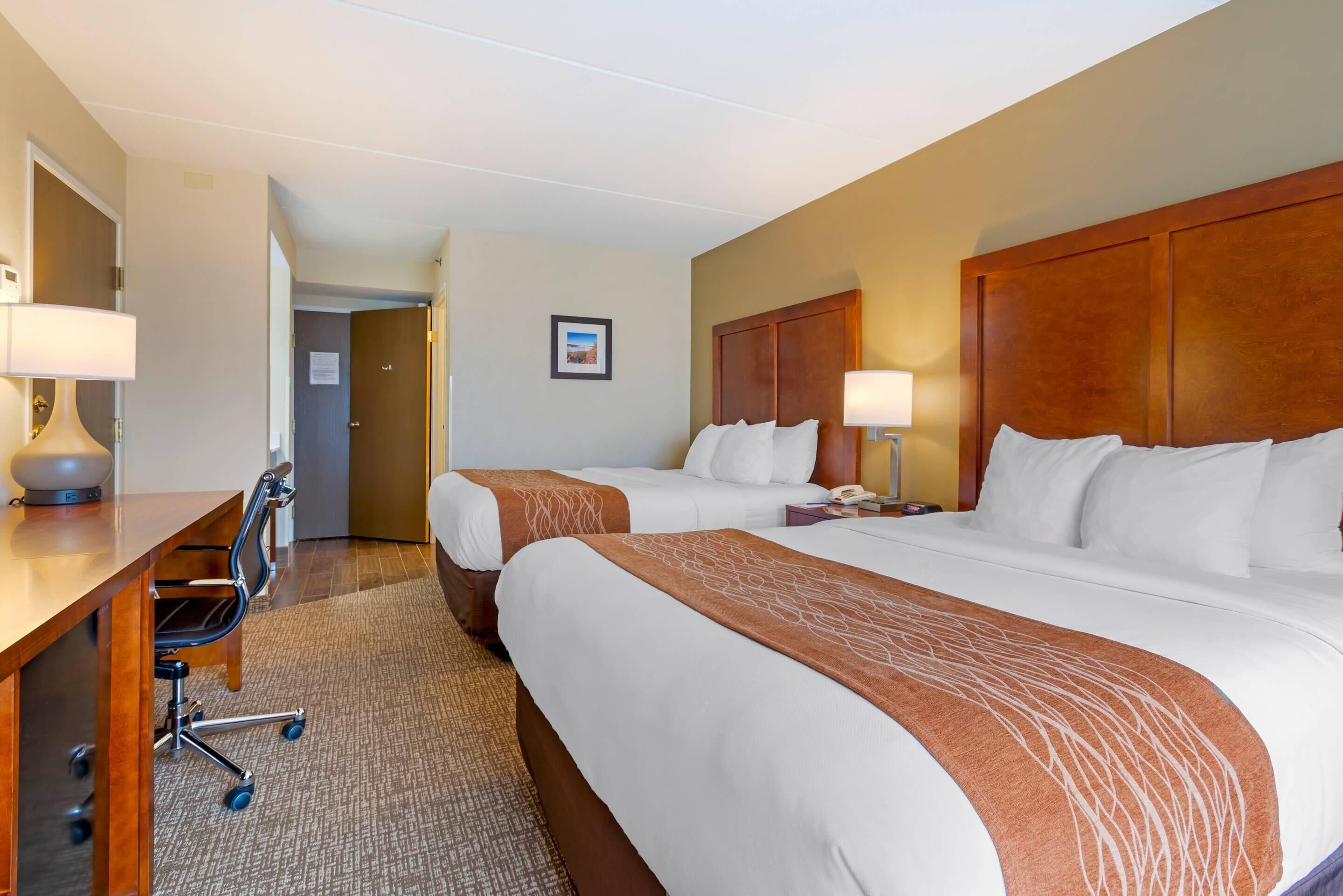 A hotel room with two beds, a desk with a chair, and a door leading to another room.