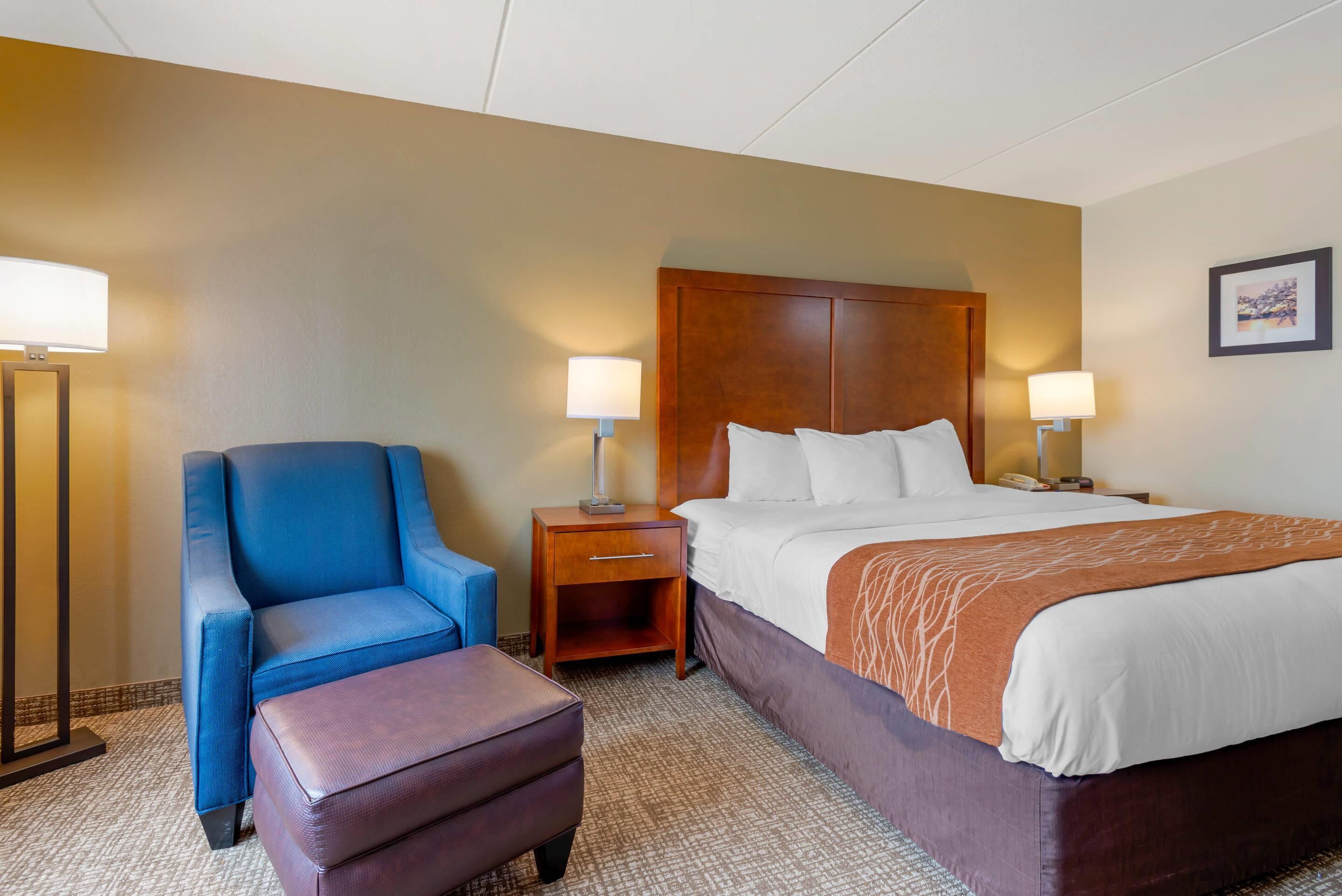 A hotel room with a bed, a blue armchair, a wooden nightstand, and a floor lamp.