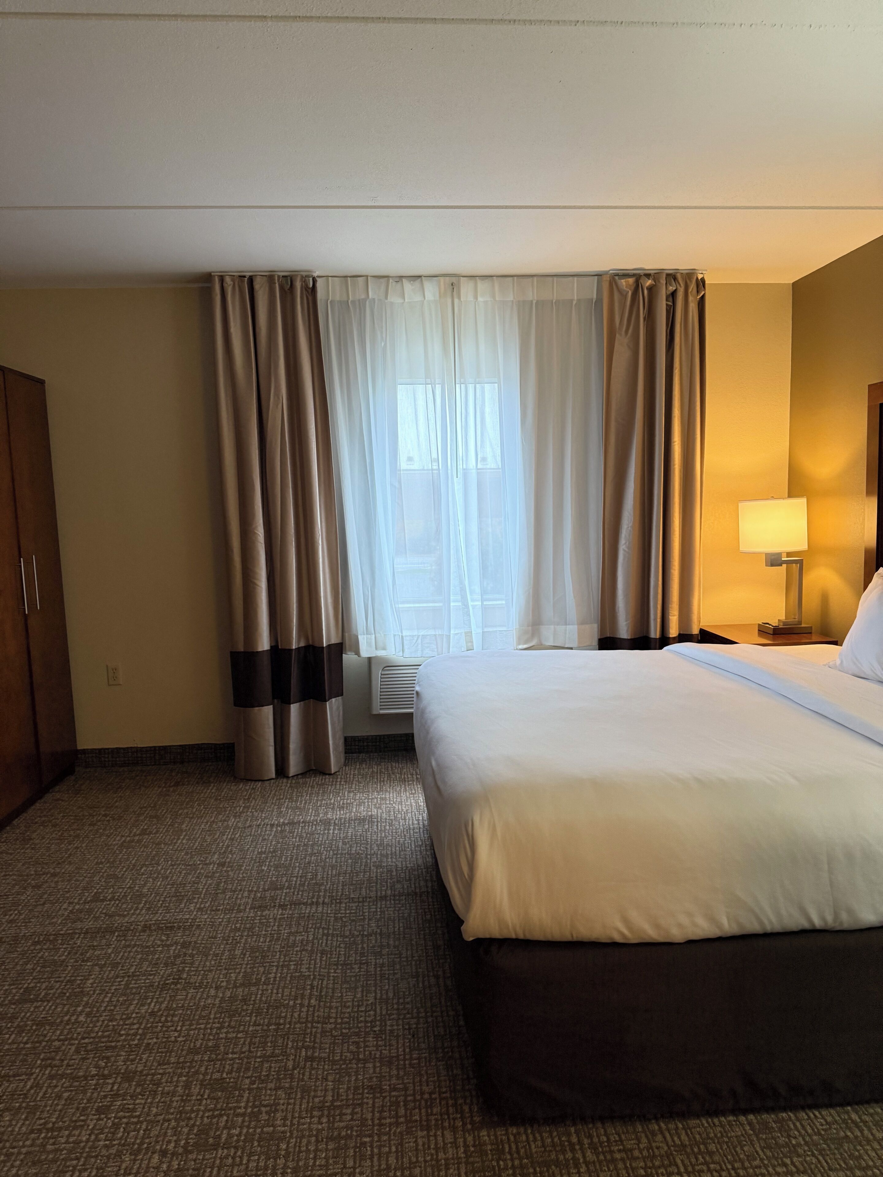 A hotel room with a large bed, a bedside table with a lamp, a wardrobe, and a window with curtains.
