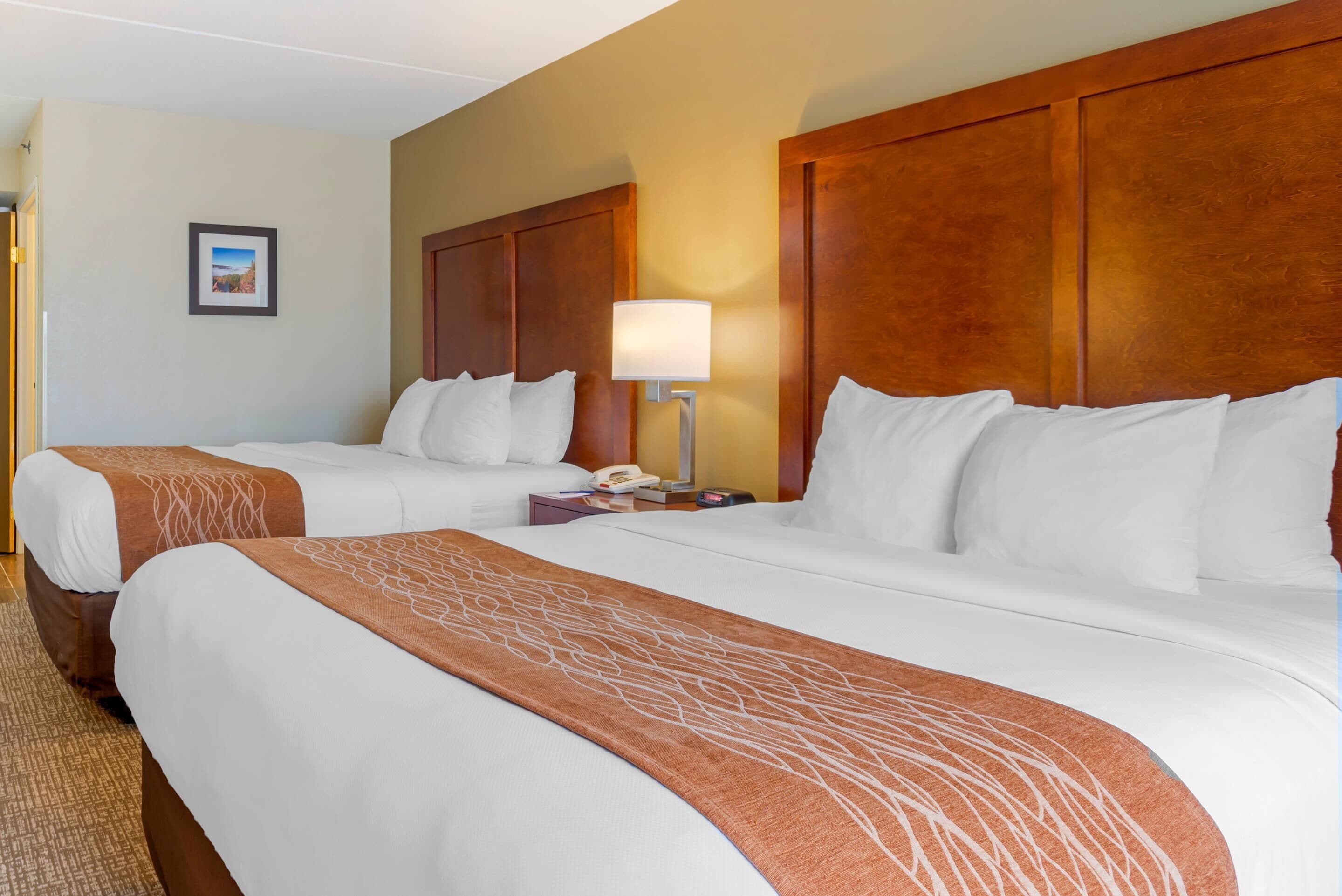 A hotel room with two beds, a wooden headboard, a bedside lamp, a phone, and a framed picture on the wall.