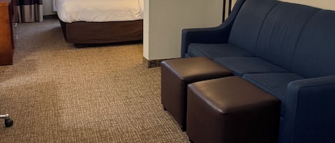 Una habitación de hotel con cama, sofá azul, puf, escritorio y lámpara.