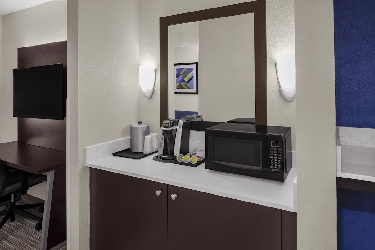 Ein Hotelzimmer mit einer Küchenzeile, darunter eine Mikrowelle, eine Kaffeemaschine und ein kleiner Kühlschrank. Es gibt einen Schreibtisch mit Stuhl sowie einen an der Wand montierten Flachbildfernseher.