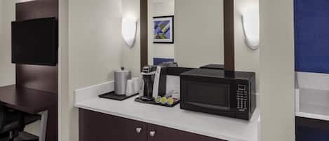 Ein Hotelzimmer mit einer Küchenzeile, darunter eine Mikrowelle, eine Kaffeemaschine und ein kleiner Kühlschrank. Es gibt einen Schreibtisch mit Stuhl sowie einen an der Wand montierten Flachbildfernseher.