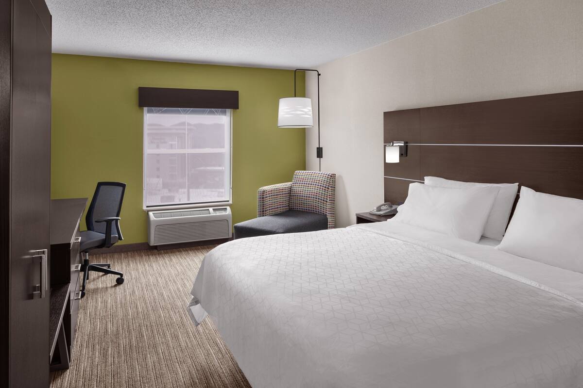 Ein Hotelzimmer mit einem großen Bett, einem Schreibtisch mit Stuhl, einem kleinen Sessel und einem Fenster mit Jalousien.