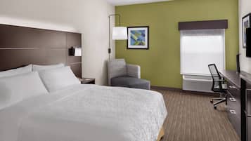 A hotel room with a large bed, a desk with a chair, a small armchair, a wall-mounted TV, and a framed picture on the wall.