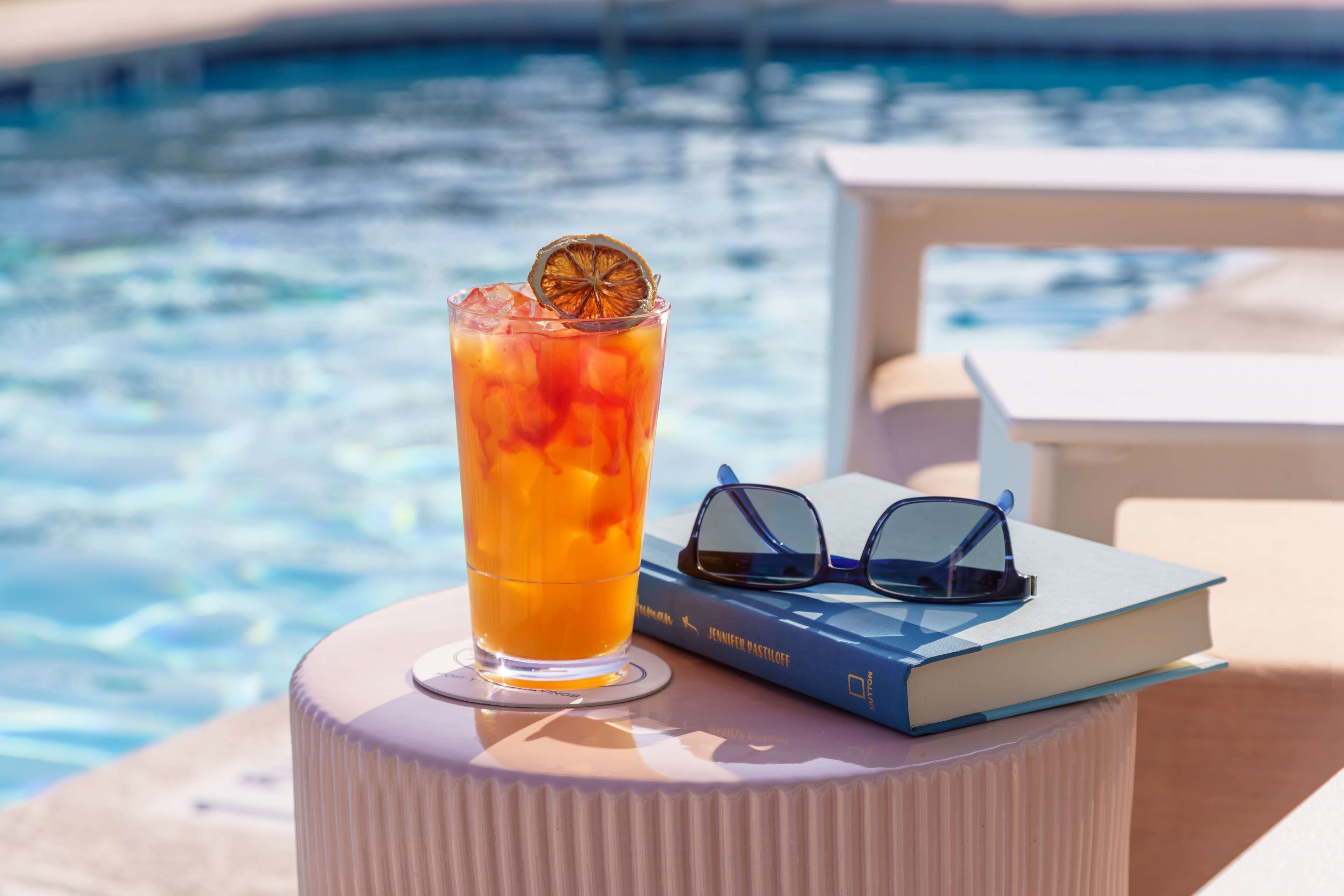 Un vaso de té helado con una rodaja de fruta sobre una mesa blanca junto a una piscina, un par de gafas de sol y un libro titulado 