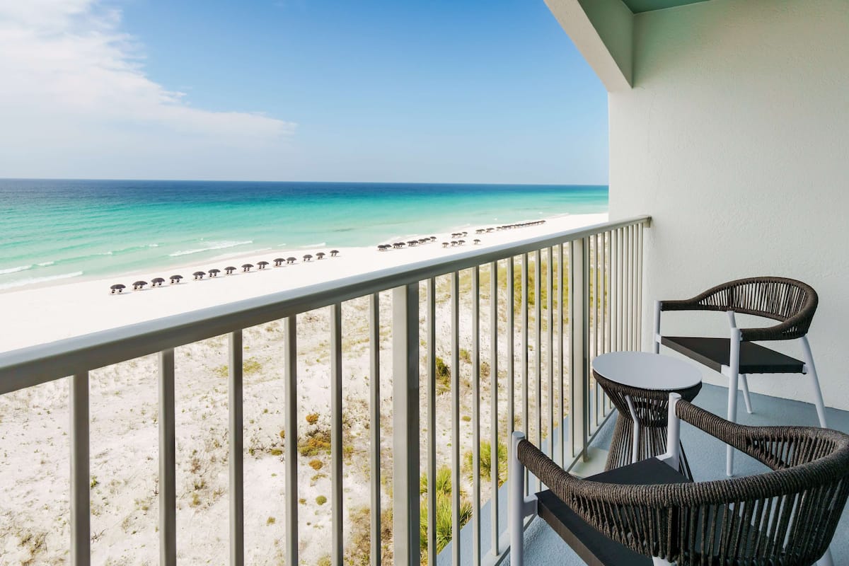 A balcony with wicker chairs and a table overlooking a beach with umbrellas.