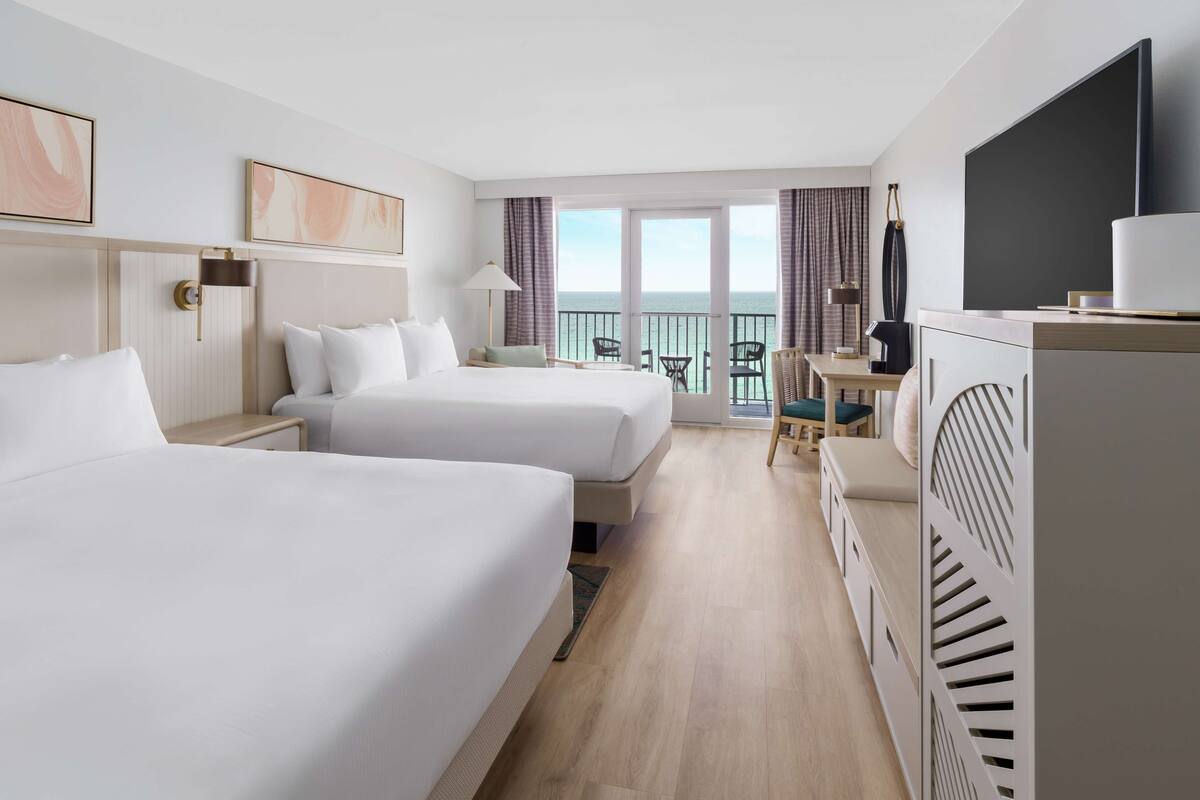 A hotel room with two beds, a desk, a TV, and a balcony with a view of the sea.