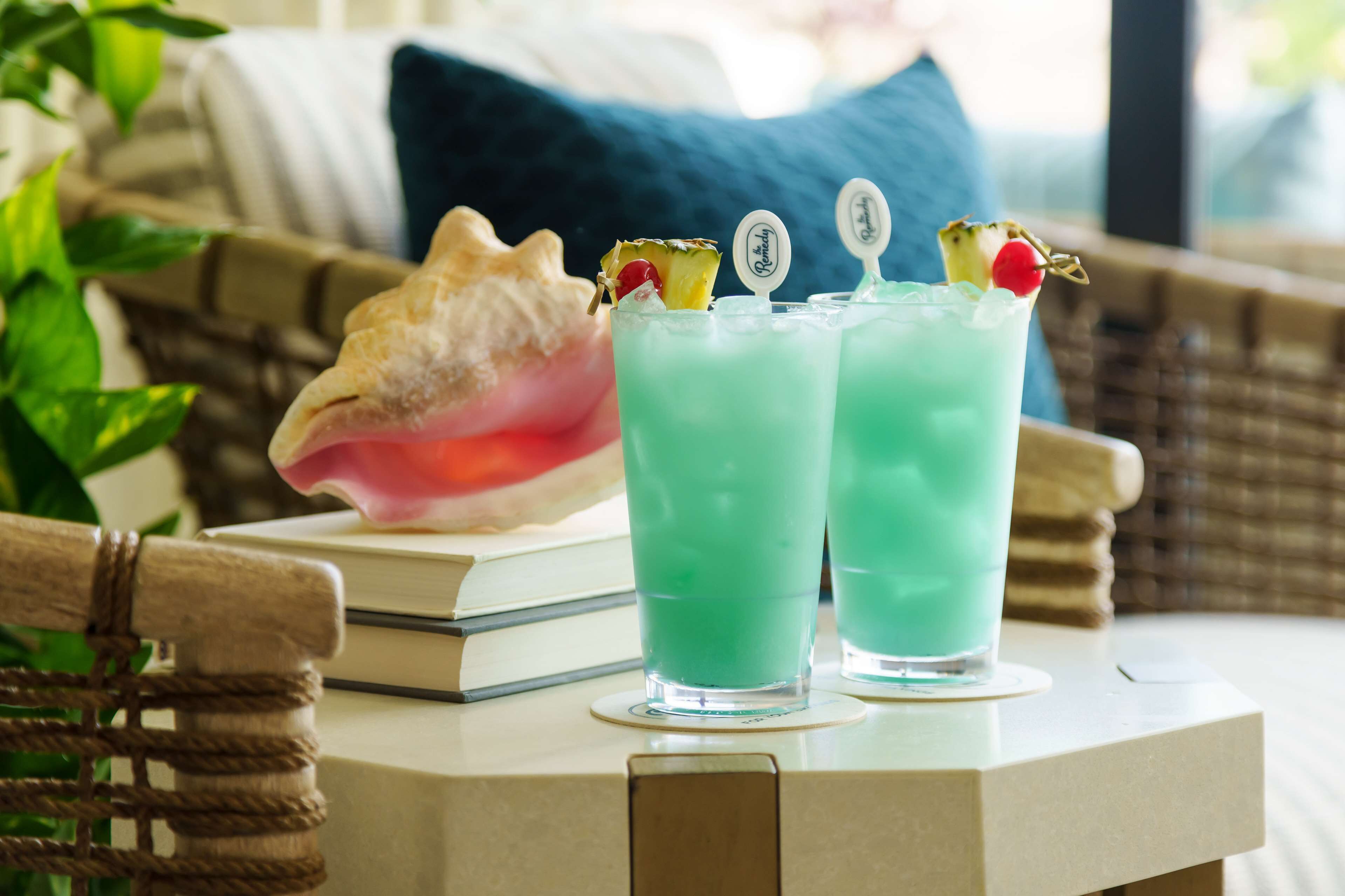 Two glasses of a green beverage with garnishes on a table, a book, and a shell in the background.