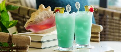 Dos vasos de una bebida verde con adornos sobre una mesa, un libro y una concha al fondo.