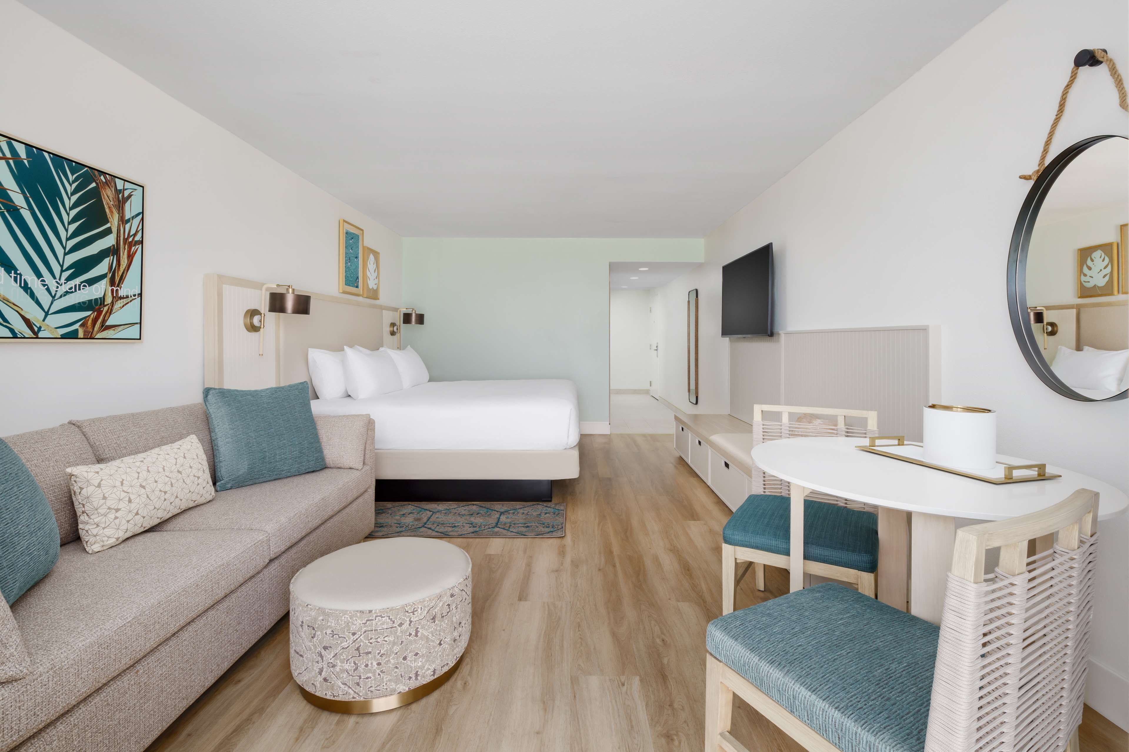 A modern hotel room with a bed, a sofa, a round ottoman, a dining table with chairs, and a large mirror.