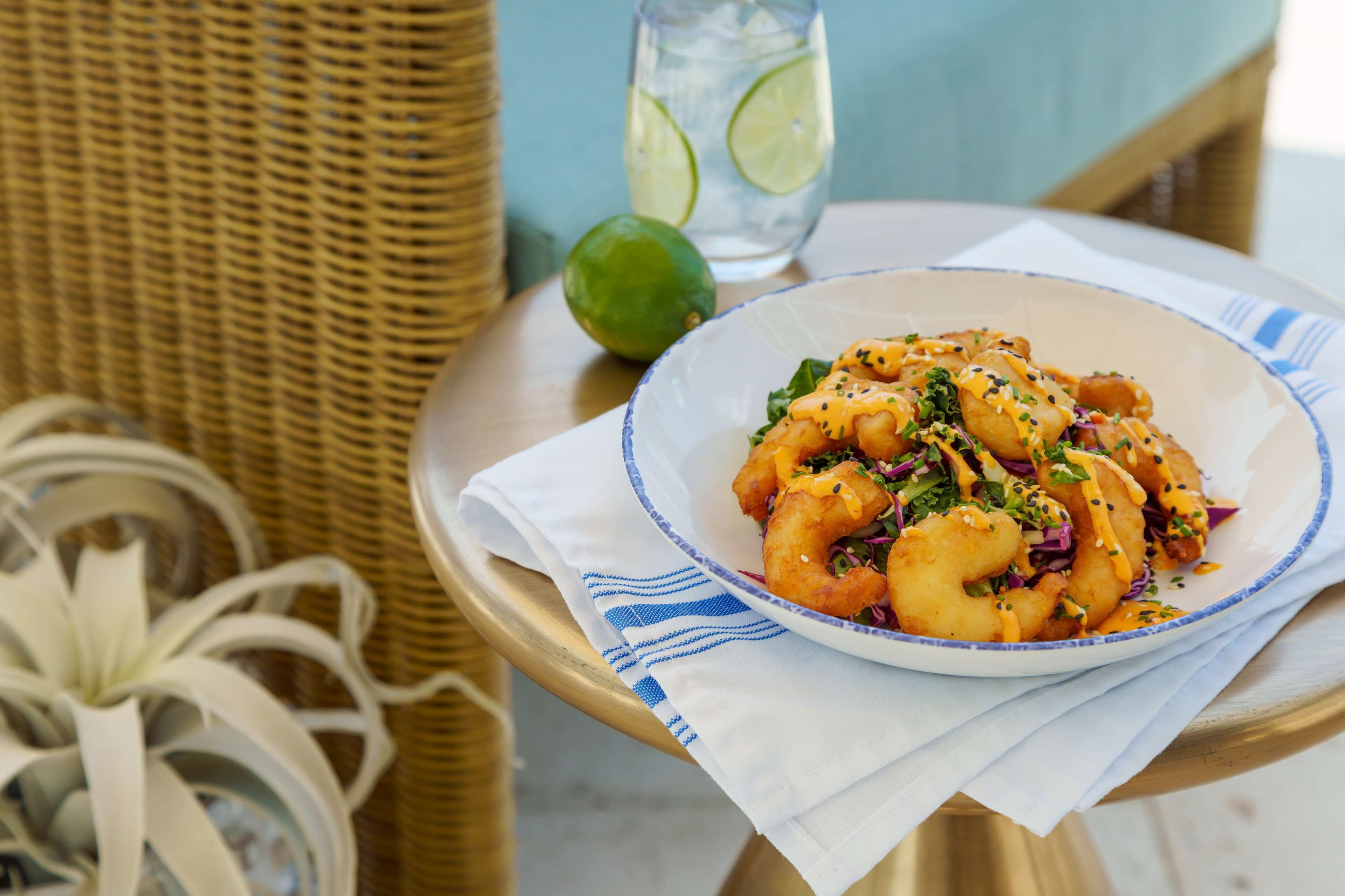 A plate of food on a table with a wicker chair and a glass of water with lime slices.