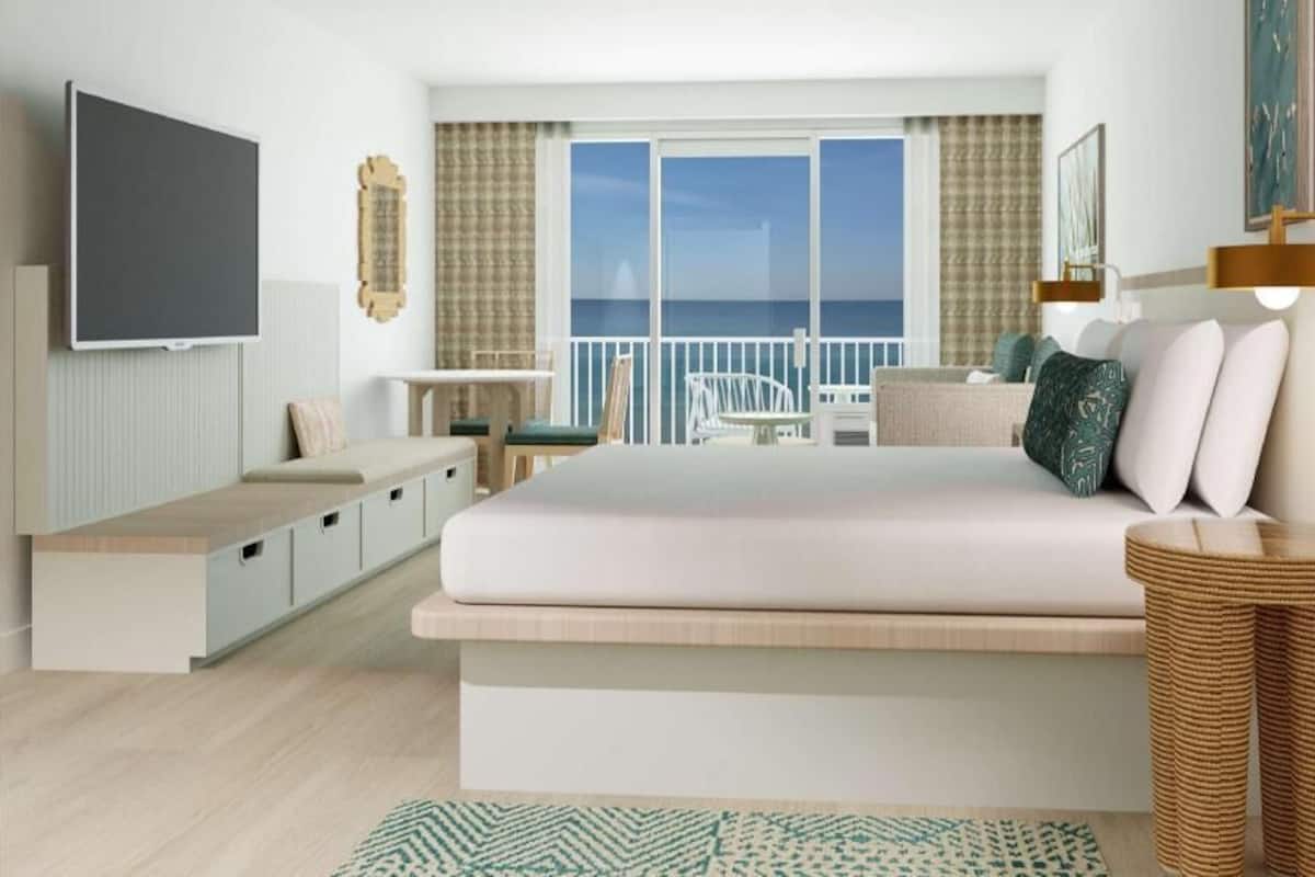 Un dormitorio moderno con una cama grande, televisión, balcón con vistas al mar y una pequeña zona de escritorio.