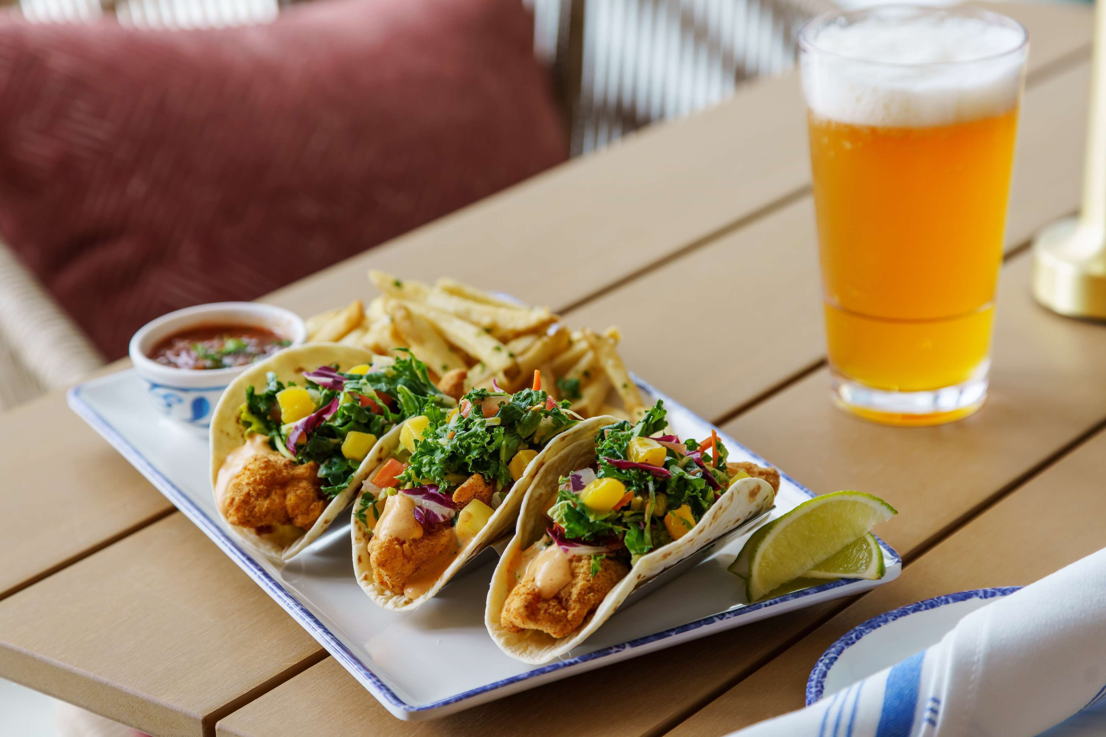 Tre tacos accompagnati da salsa, un bicchiere di birra e una fetta di lime su un tavolo di legno.