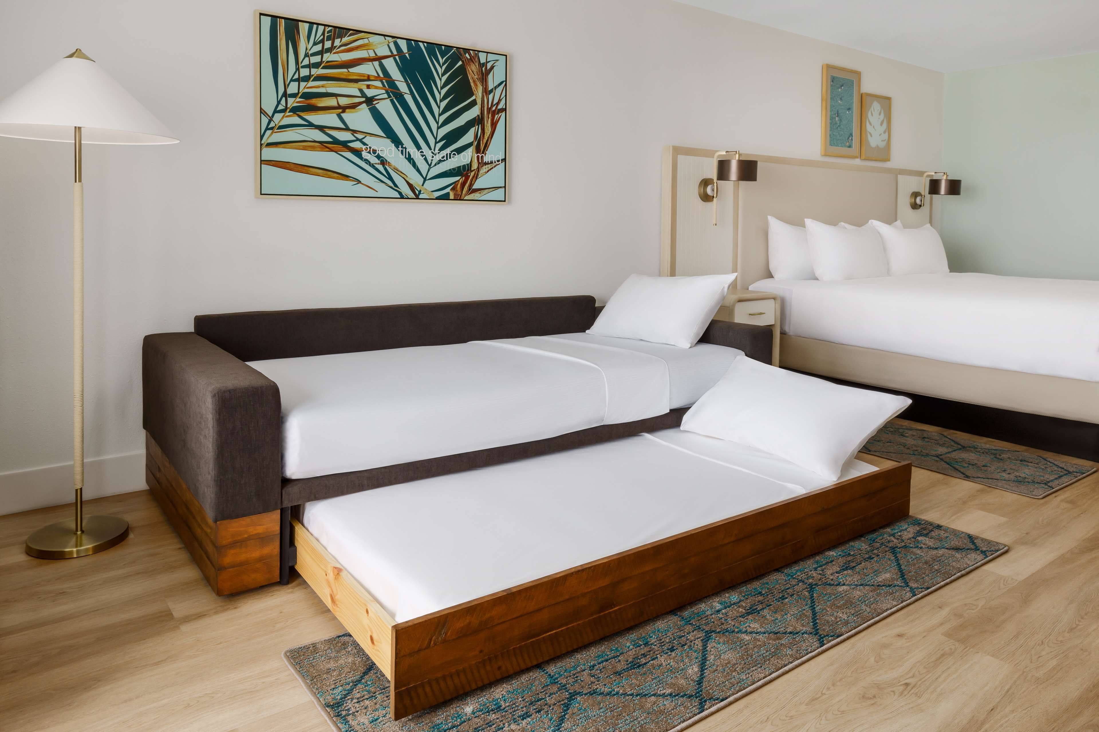 A hotel room with a sofa bed, a single bed, and a bedside table with a lamp.