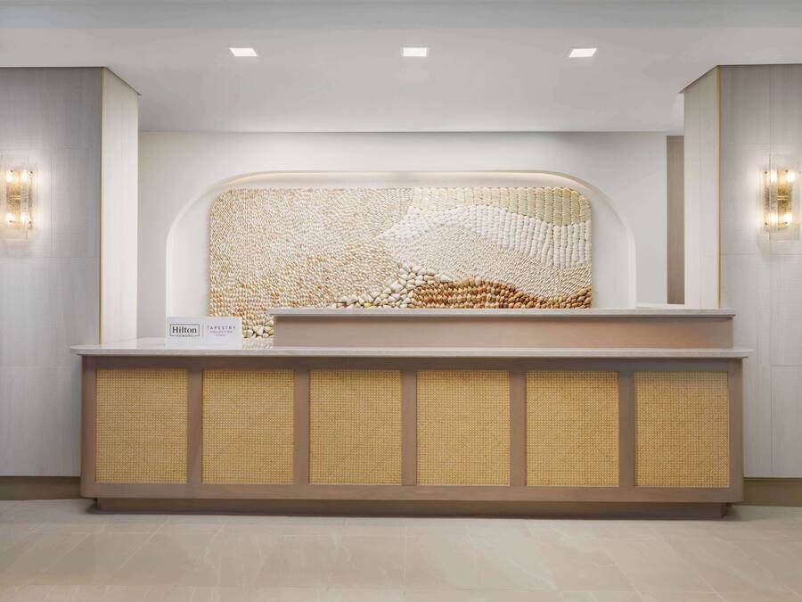A reception desk with a woven tapestry backdrop, a sign indicating a Hilton hotel, and two wall-mounted light fixtures.