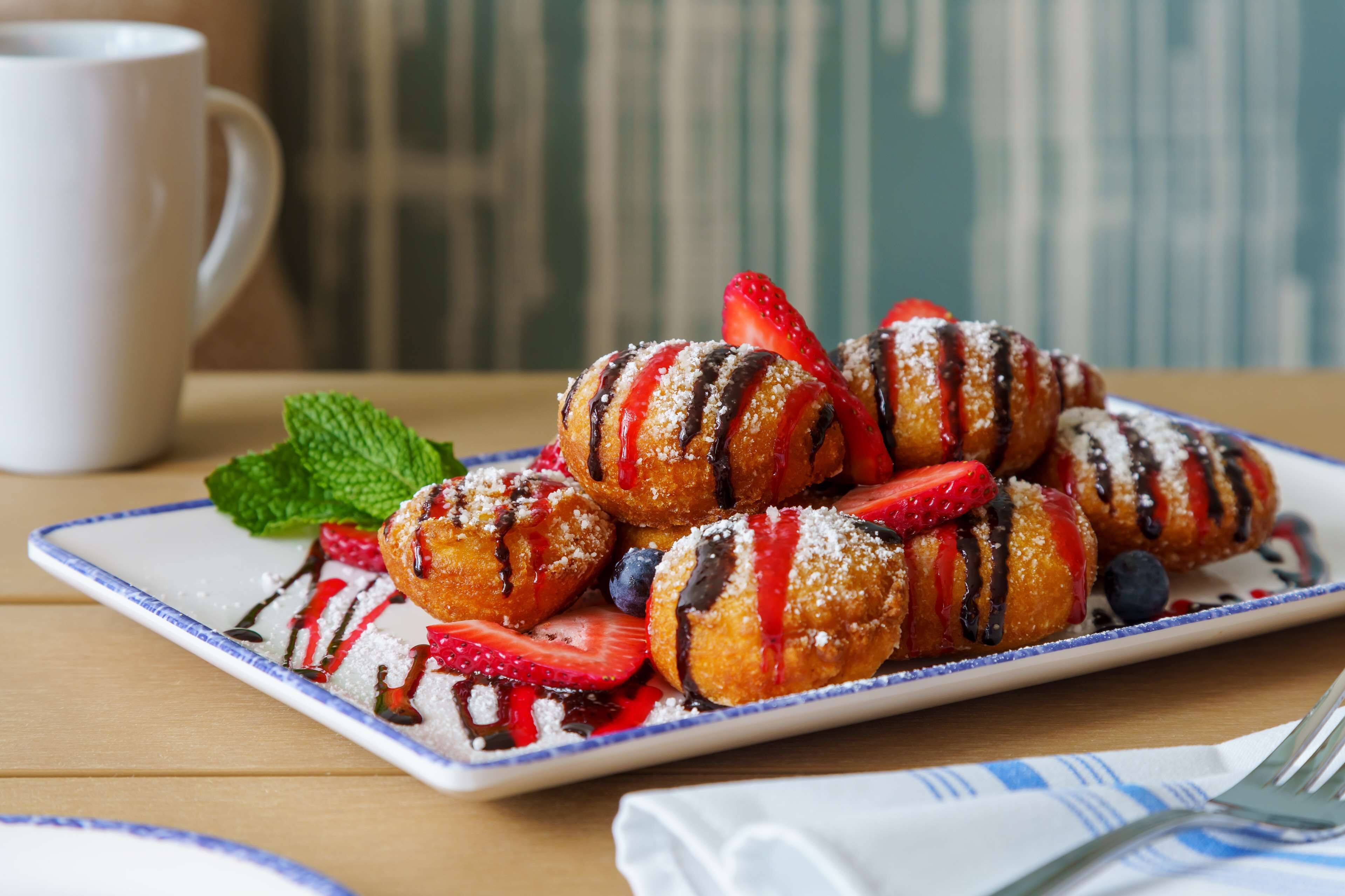 A plate of pastries with strawberries and blueberries, a white mug, and a napkin with a fork on a wooden table.