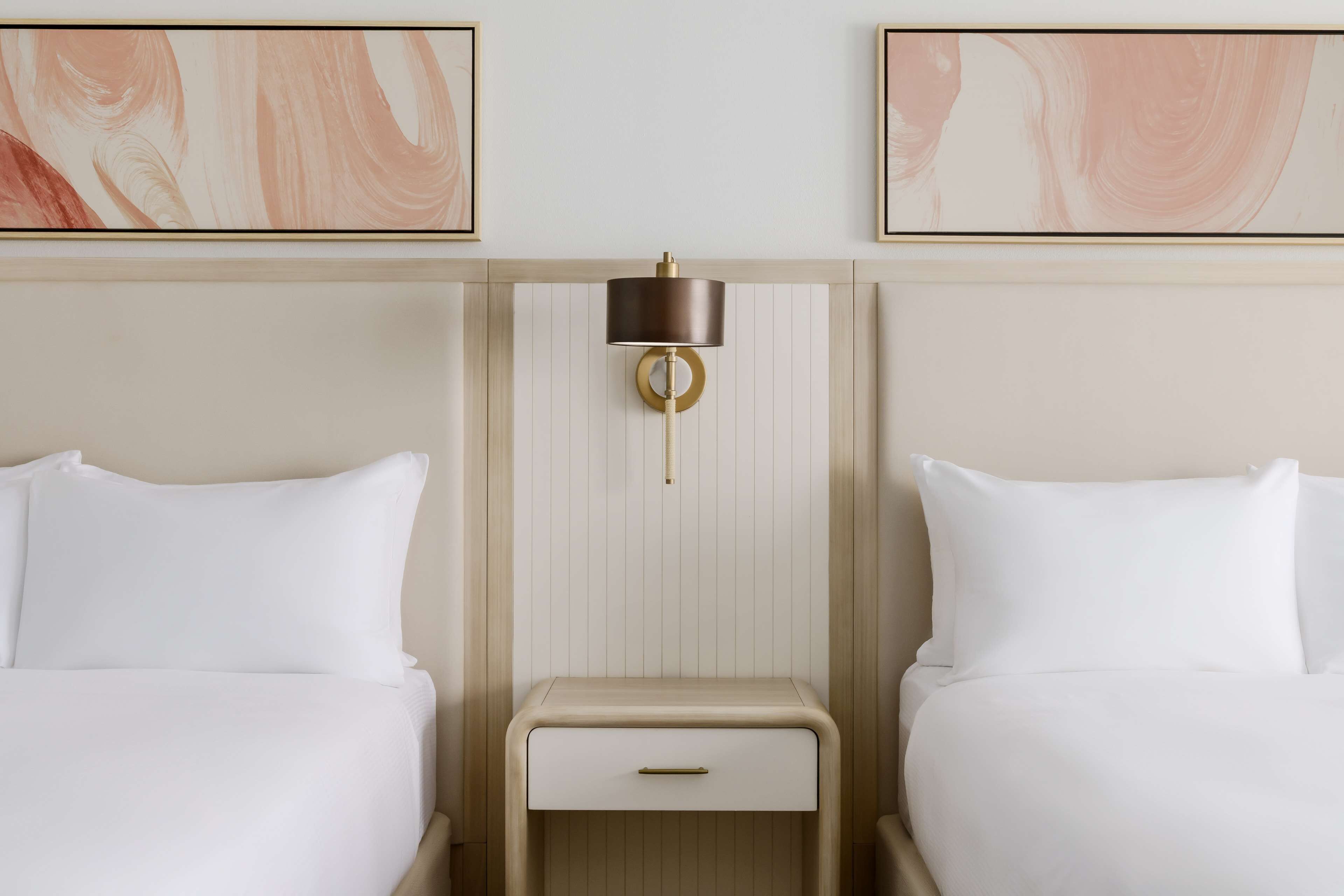 A hotel room with two beds, a bedside table with a lamp, and abstract artwork on the walls.