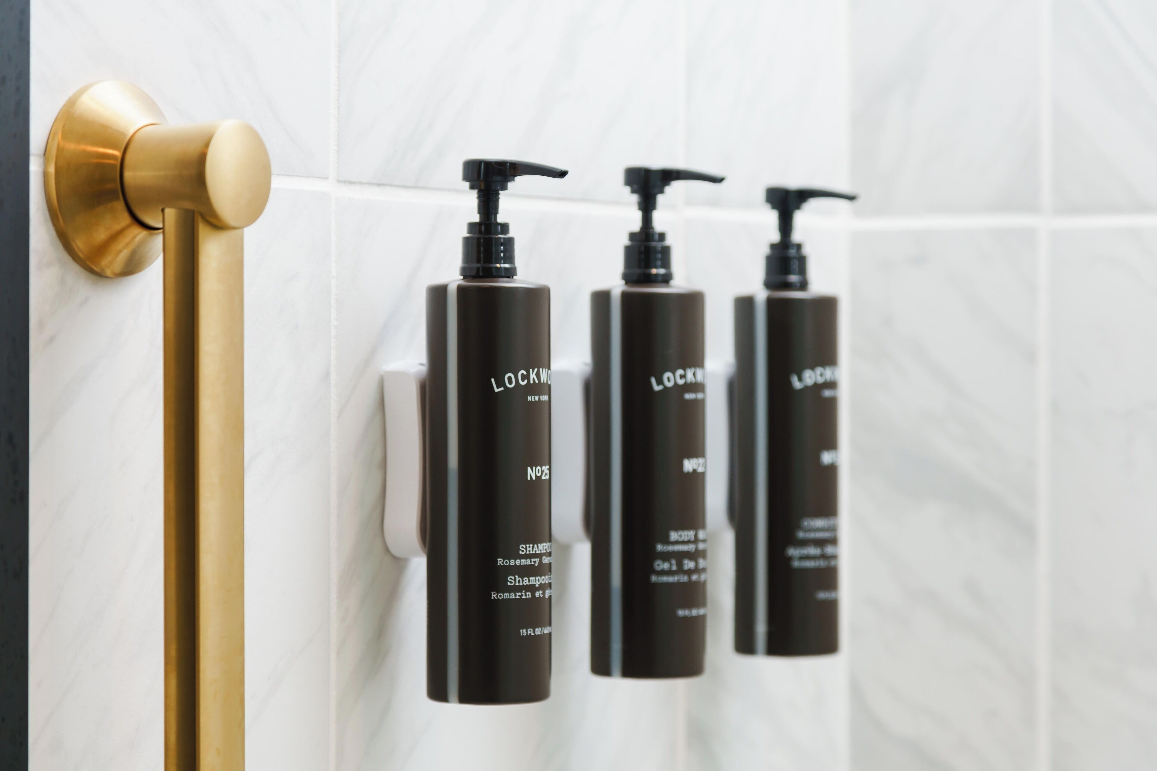 A bathroom with a gold towel bar, three black bottles of Lockwood body wash, and a white tiled wall.