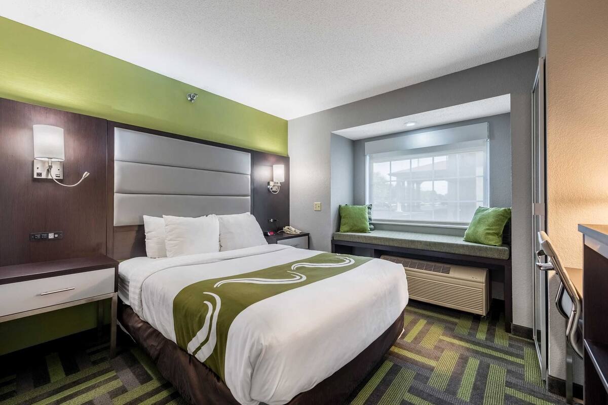 A hotel room with a bed, bedside table, lamp, window seat, and a view of the outside.