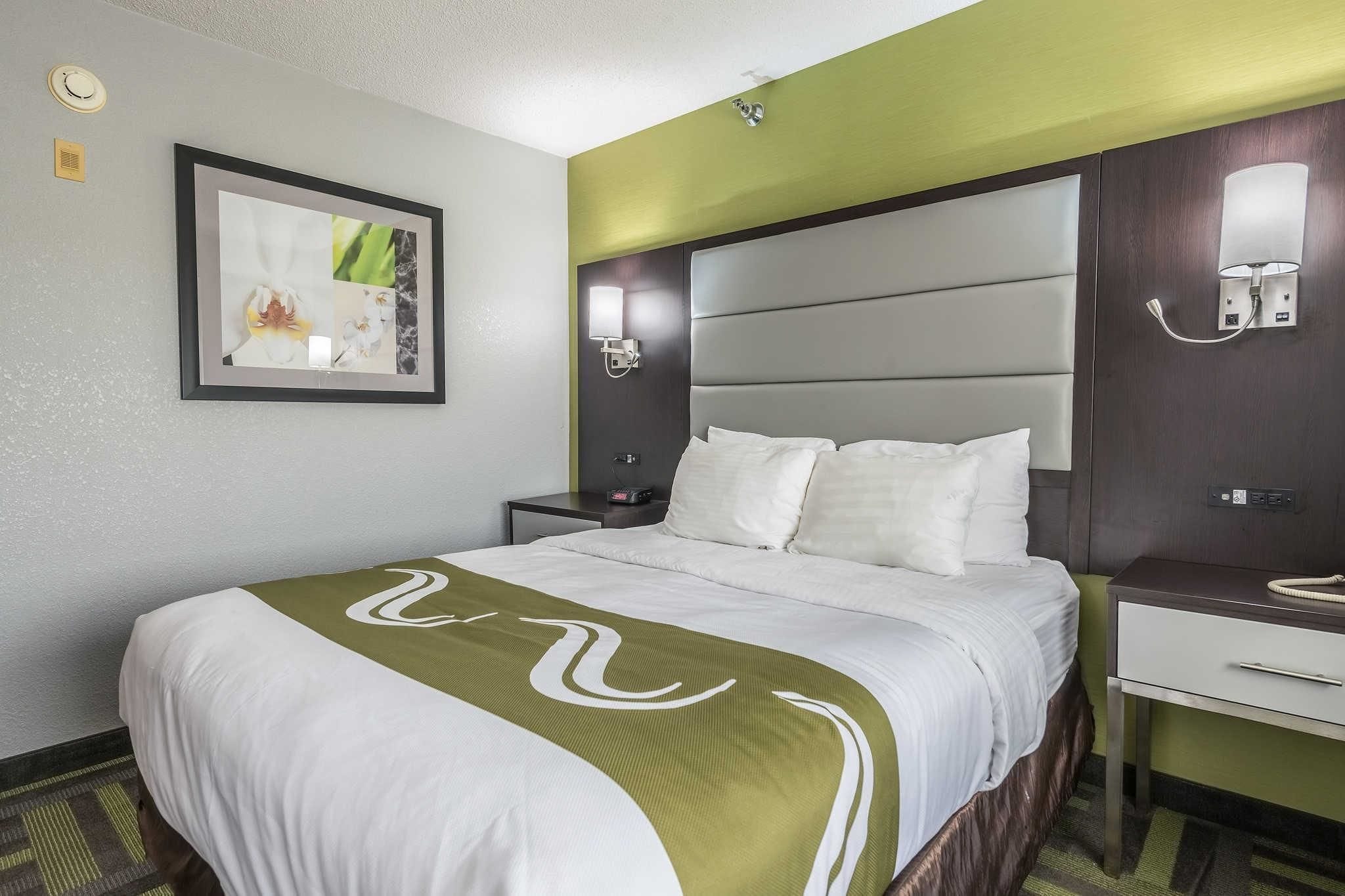 Een hotelkamer met een groot bed, een nachtkastje, een lamp aan de muur en een schilderij in een lijst.
