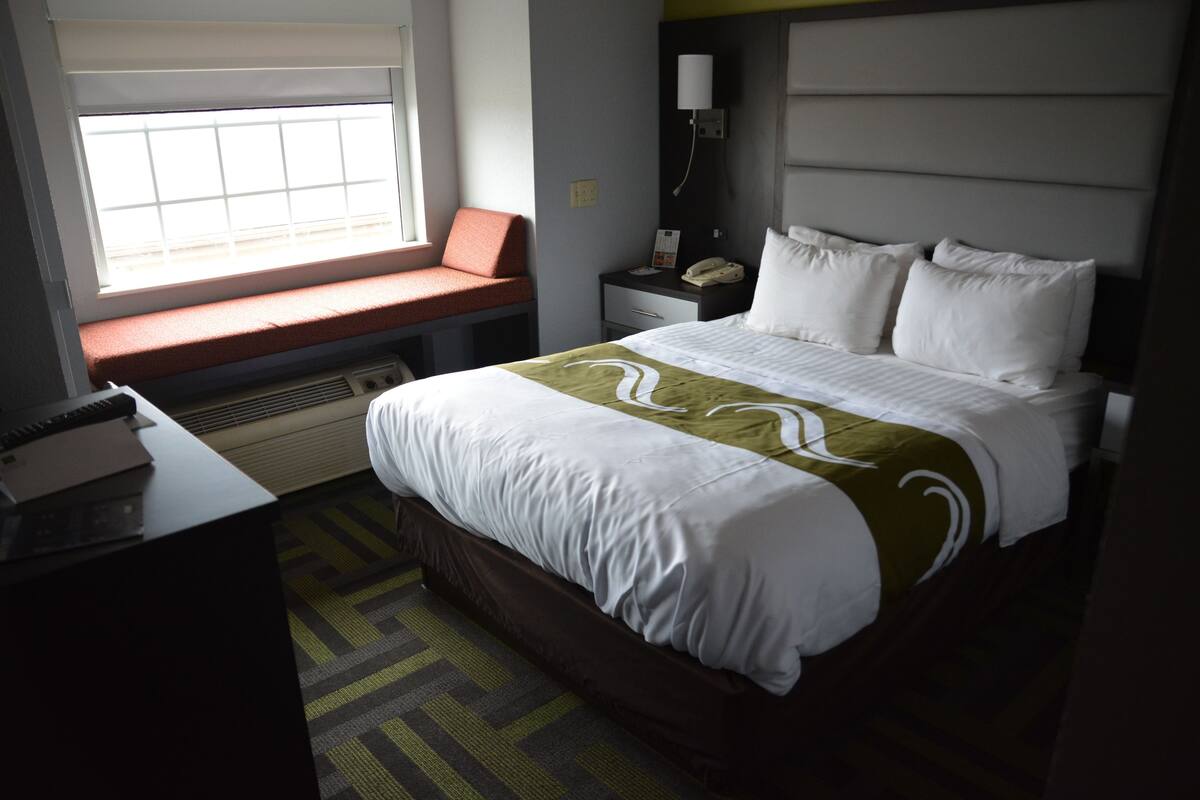 A hotel room with a bed, a desk, a window seat, and a nightstand with a lamp.