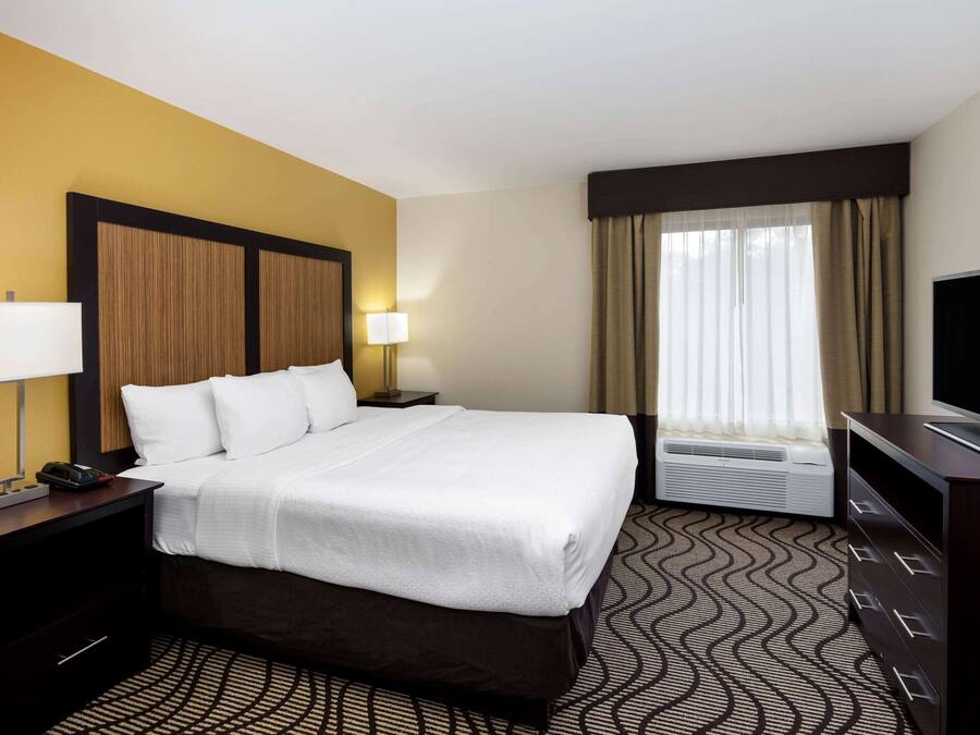 A hotel room with a large bed, a bedside table with a lamp, a television, and a window with curtains.