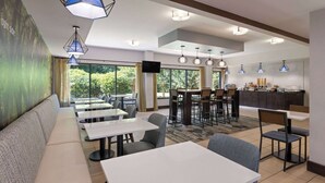 Un restaurante moderno con una barra larga, varias zonas de estar y amplios ventanales con vistas a zonas verdes.