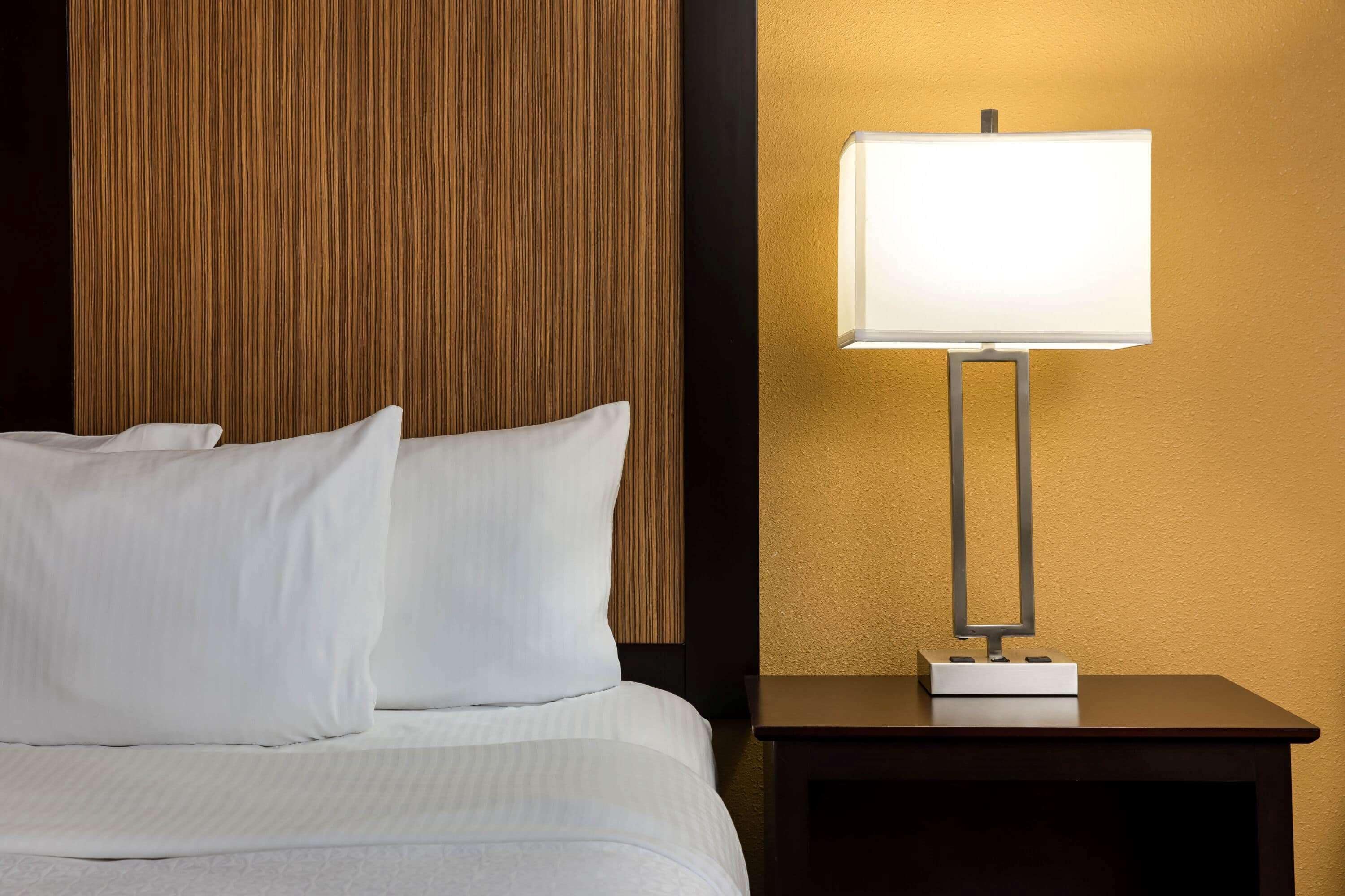 A neatly made bed with white linens and pillows, a wooden headboard, and a bedside table with a lamp.