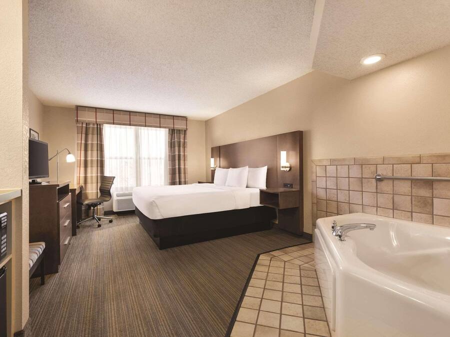 A hotel room with a large bed, a separate desk with a chair, a desk lamp, a flat-screen TV, a microwave, a built-in oven, a large bathtub, and a window with curtains.