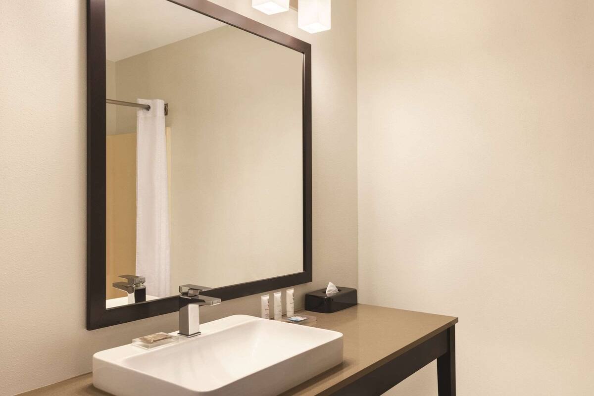 Ein modernes Badezimmer mit einem großen Spiegel, einem weißen Waschbecken und einer kleinen, schwarzen Ablage mit Toilettenartikeln.