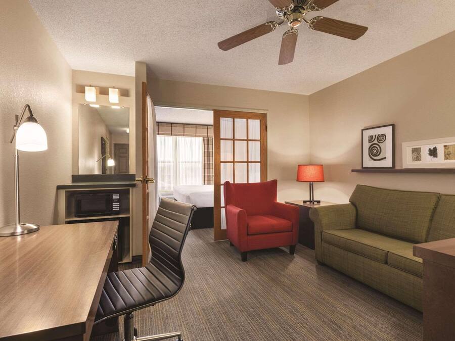 Ein Hotelzimmer mit einem Holztisch, einem roten Sessel, einem grünen Sofa, einem Deckenventilator und einem Bett mit weißer Bettdecke.