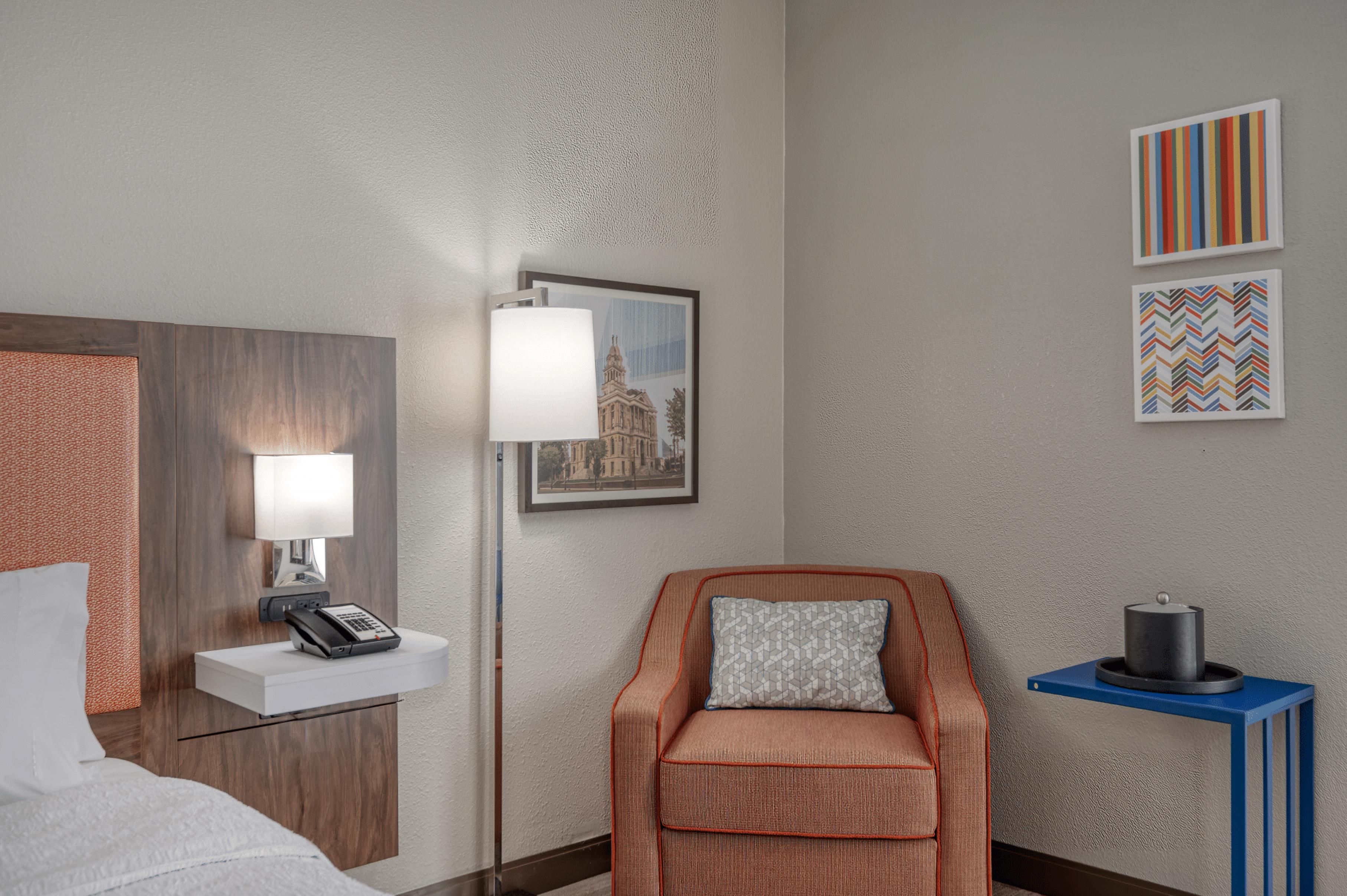 A hotel room with a bed, a desk with a lamp, a chair, a small table with a container, and two framed pictures on the wall.