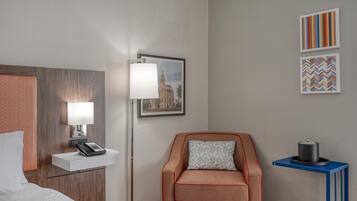 A hotel room with a bed, a desk with a lamp, a chair, a small table with a container, and two framed pictures on the wall.