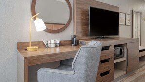 A modern hotel room with a wooden desk, a grey chair, a round mirror, a flat-screen TV, a coffee maker, and a microwave.