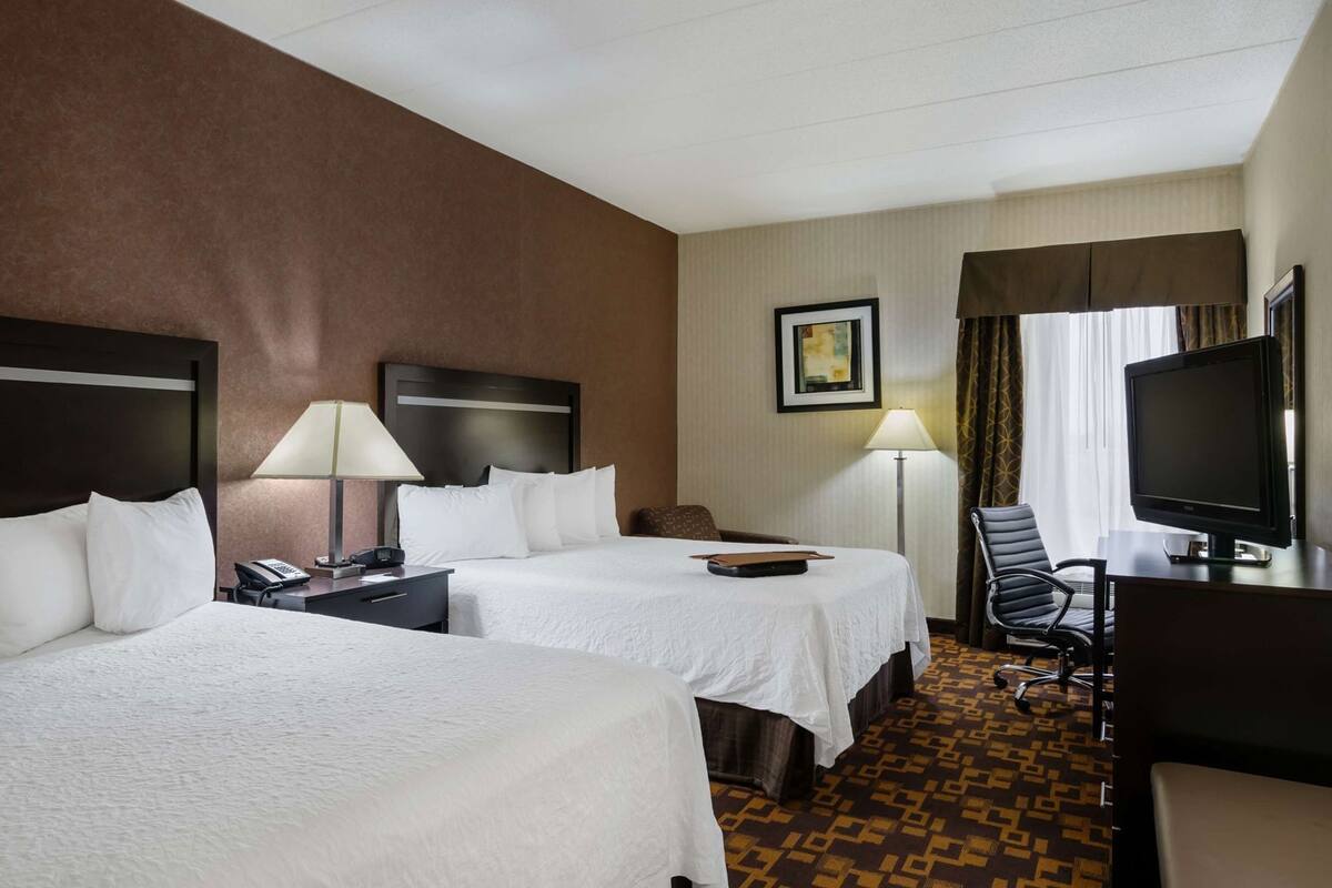 A hotel room with two beds, a desk, a chair, a television, and a lamp.