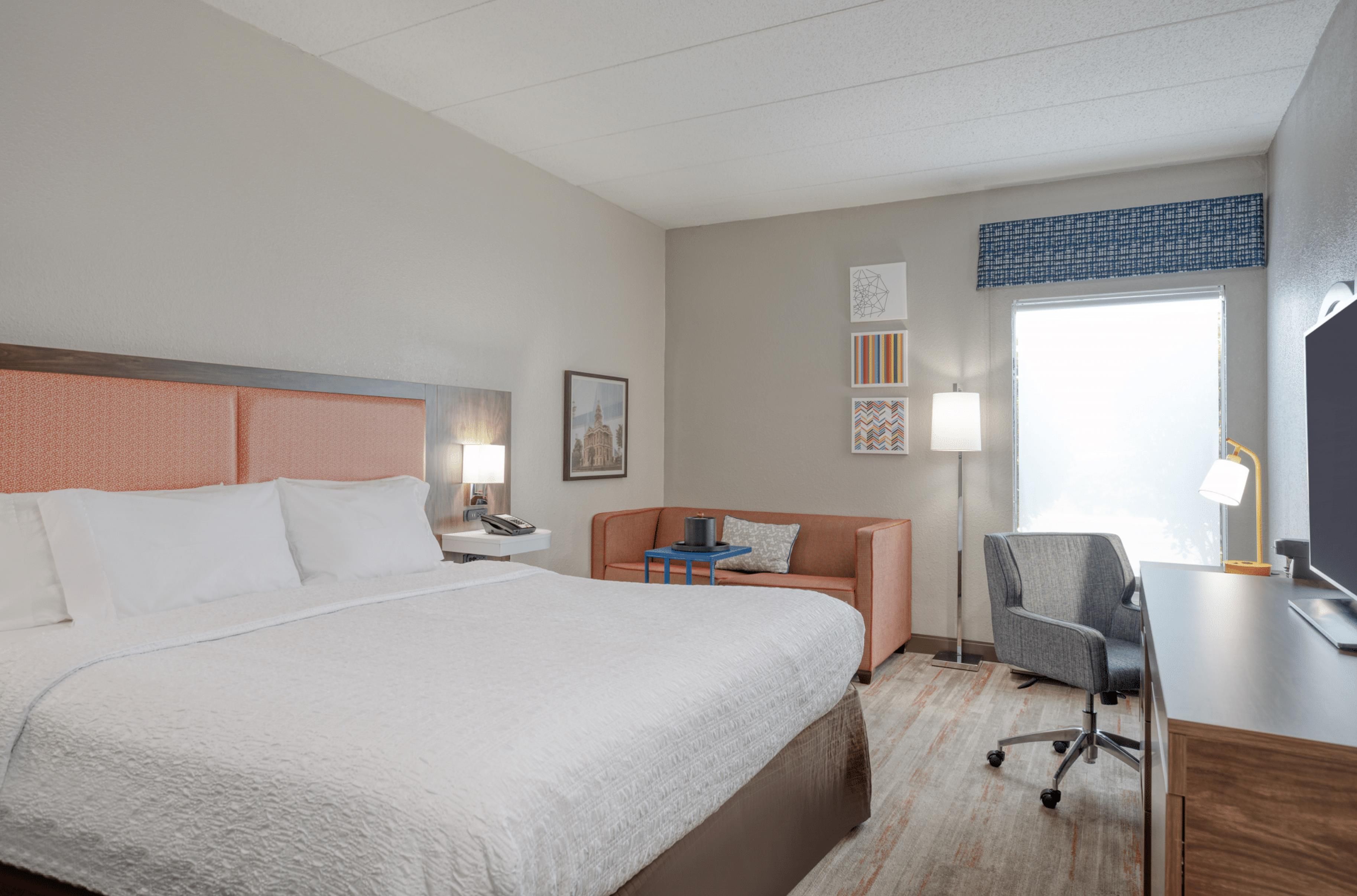 Quarto de hotel com uma cama grande, uma escrivaninha com cadeira, luminária e janela com vista.