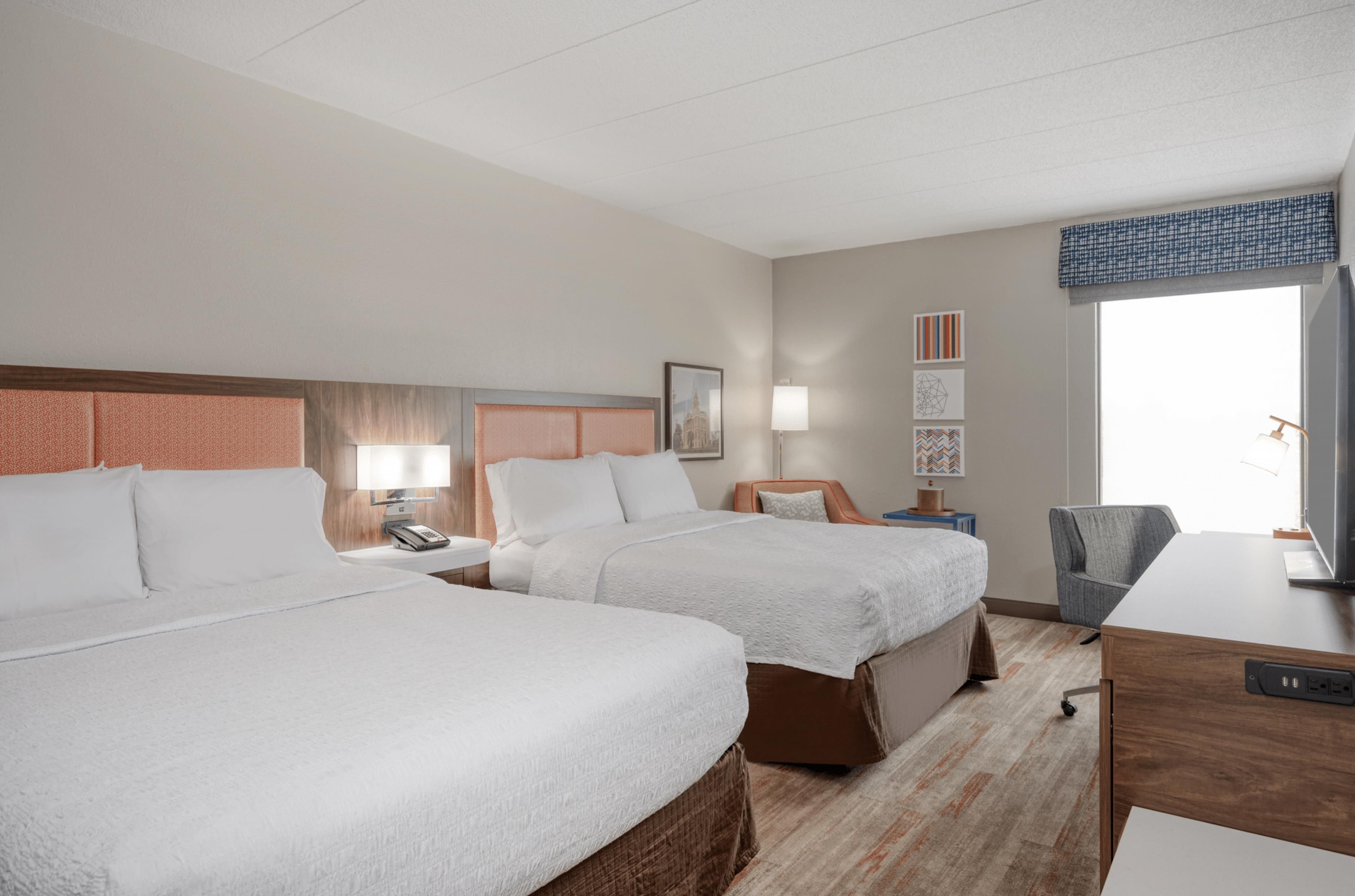 A hotel room with two beds, a desk, a chair, a TV, and artwork on the wall.