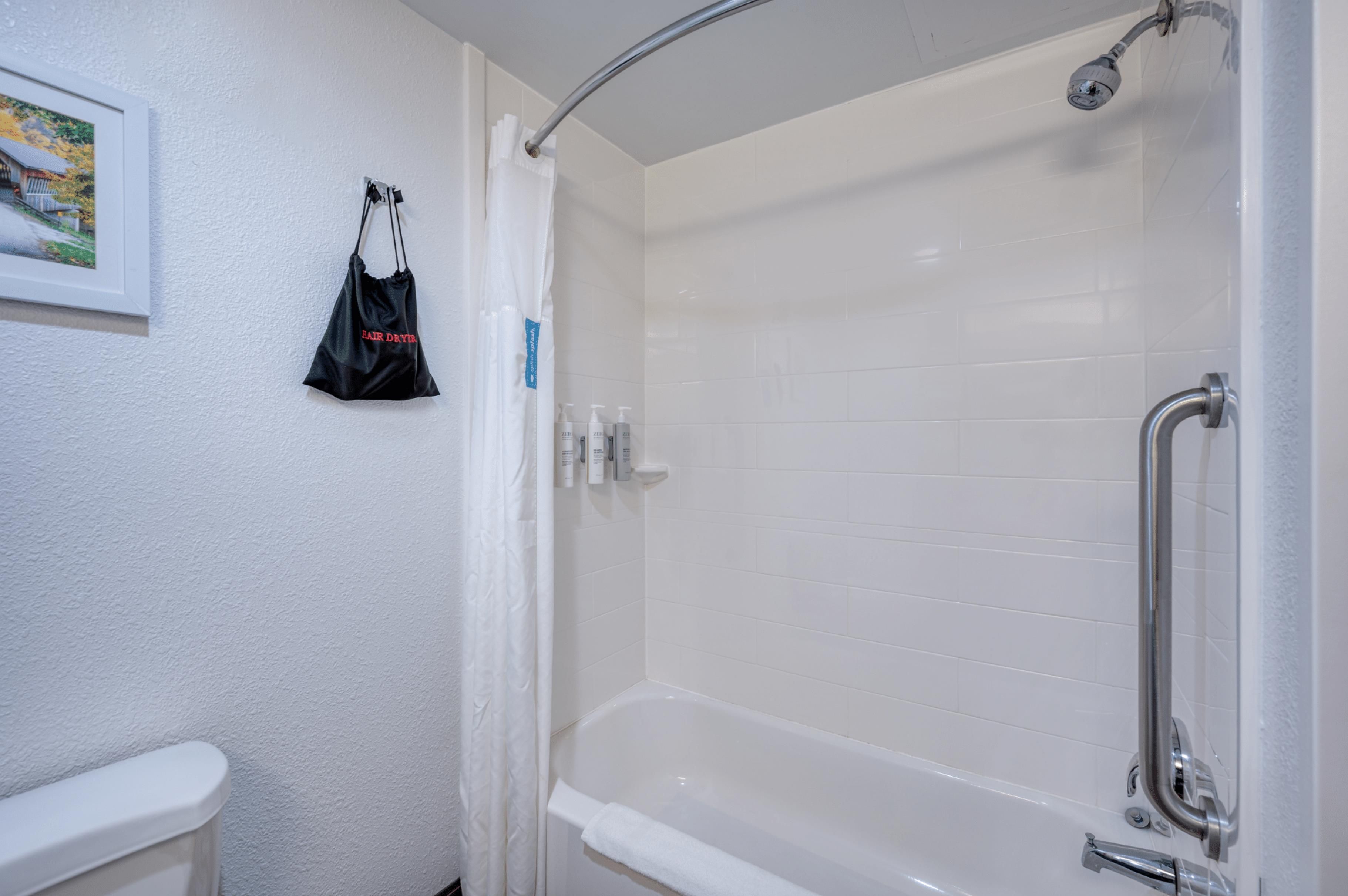 A bathroom with a white bathtub, a shower, and a toilet. There is a framed picture on the wall and a black bag hanging on the wall.
