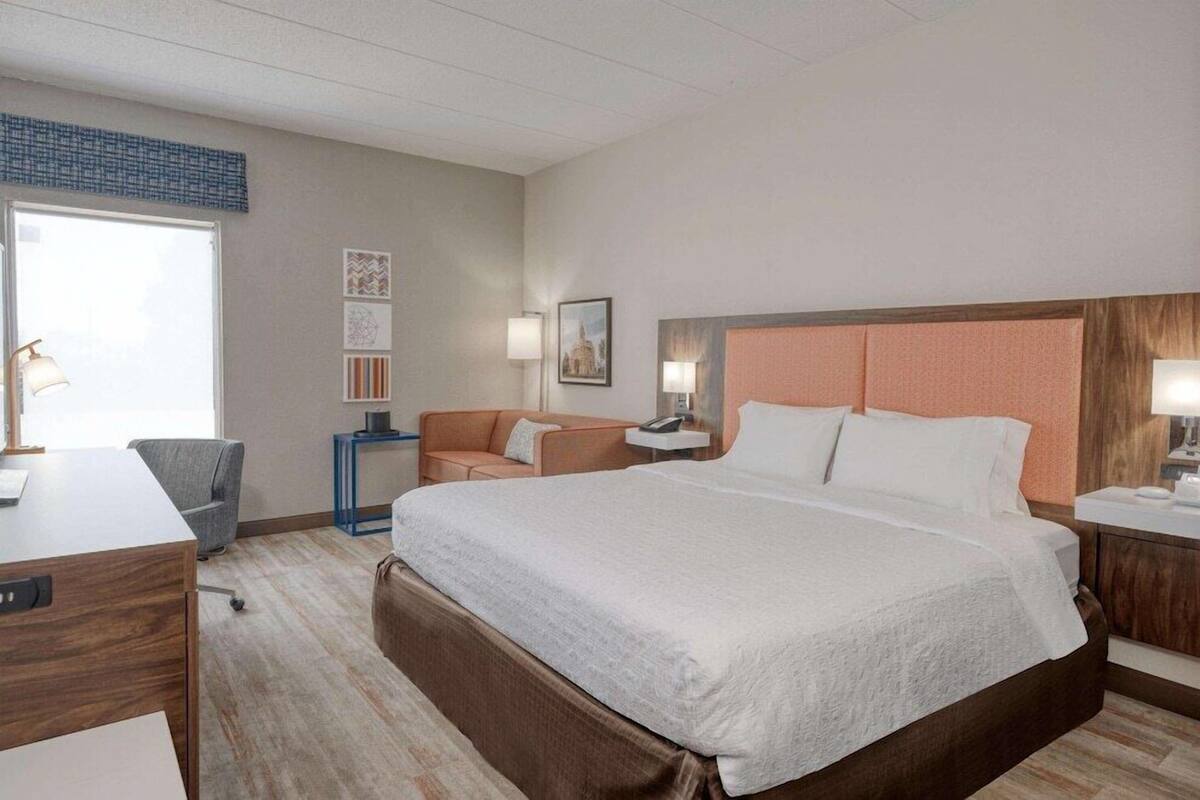 A hotel room with a large bed, a desk, a chair, and a nightstand with a lamp.