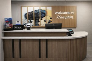 A hotel reception desk with a 'welcome to Hampton' sign, a computer, a phone, and a 'Hilton Honors' card.