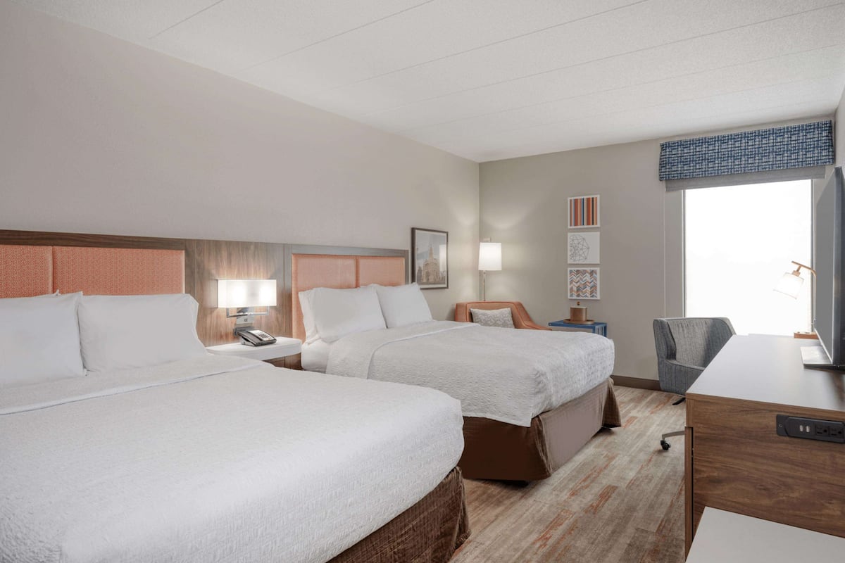A hotel room with two beds, a desk, a chair, a TV, and a window with a view.