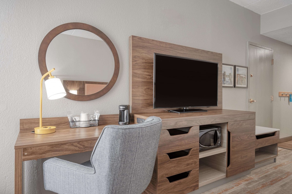 A modern hotel room with a wooden desk, a flat-screen TV, a mirror, and a grey chair.