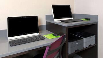 Two laptops on a desk with a keyboard, mouse, and a green mouse pad. There is a chair in the foreground and a shelf with printers in the background.