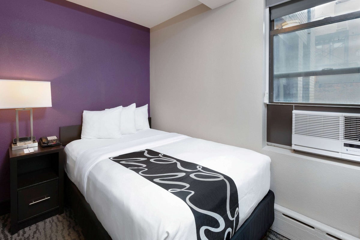 Une chambre d’hôtel avec un lit, une table de chevet, une lampe et une fenêtre avec des stores.