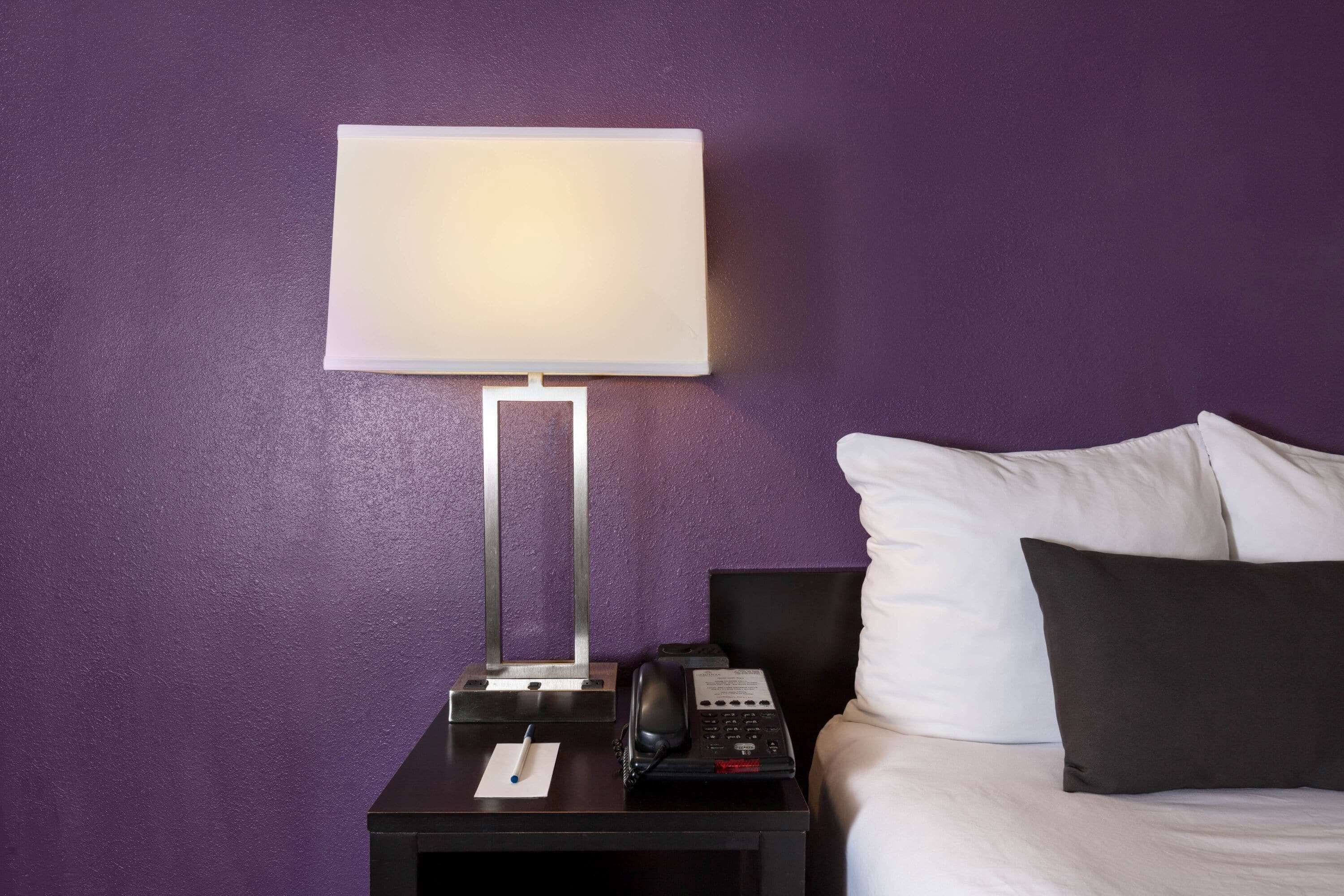 A hotel room with a bed, a nightstand with a lamp, a telephone, and a notepad.